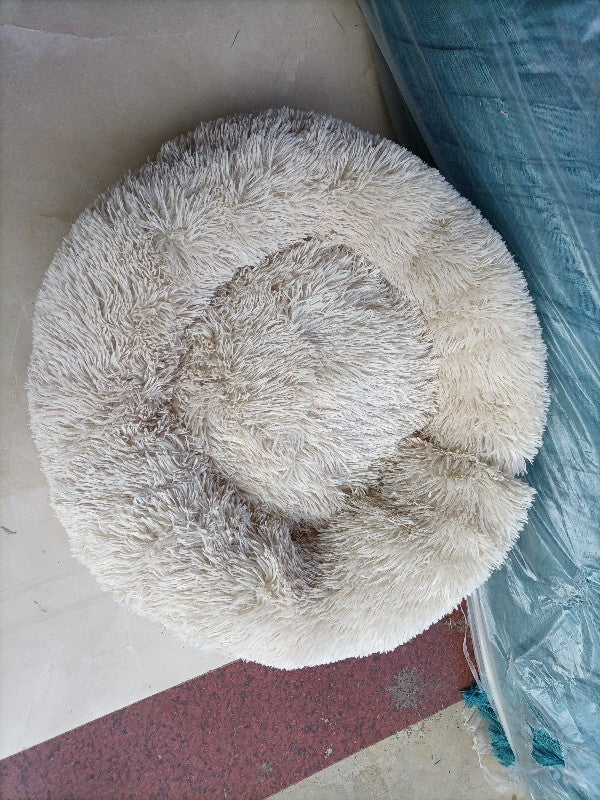 Round, fluffy pet bed on a concrete floor with a blue plastic bag in the background.